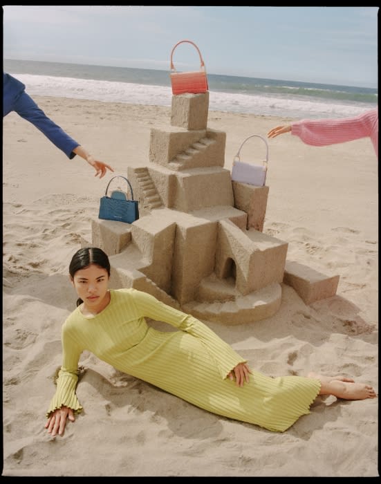 Woman in lime dress poses on beach near sandcastle with handbags; blue, pink arms reach in from sides toward handbags on the castle.
