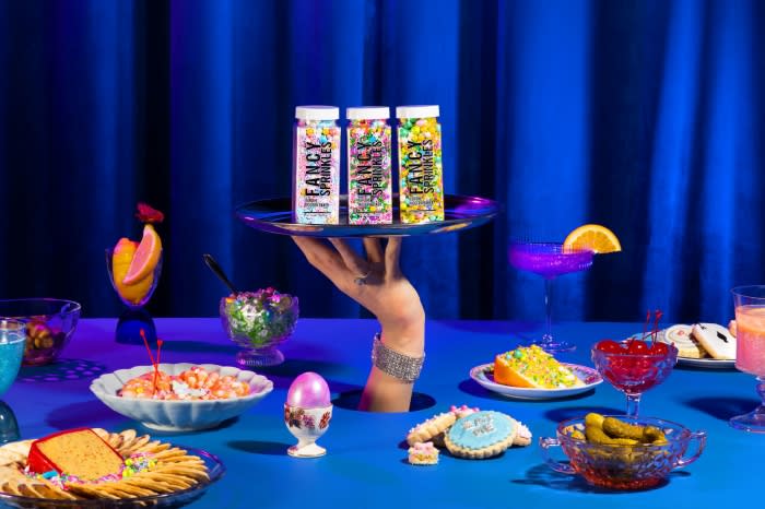 Three jars of Fancy Sprinkles sit on a silver platter held by a hand emerging from a blue table surrounded by sweets and drinks.