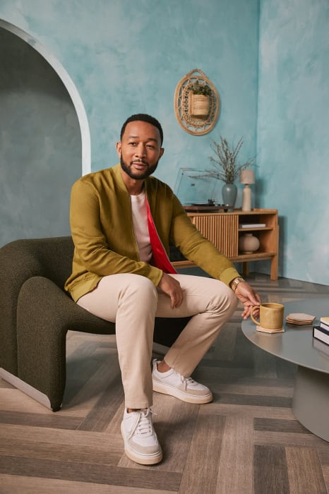 John Legend sits in a modern chair in a casual olive-green jacket, tan pants, and white shoes, holding a yellow mug indoors.