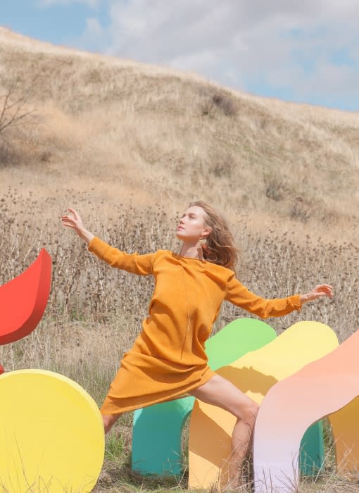A woman in a mustard dress stands amidst colorful, oversized shapes in a grassy field with a hillside under a bright blue sky.