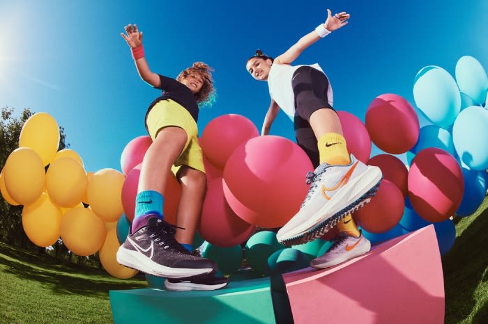 Two kids strike playful poses beside balloons and athletic shoes. Nike shoes are featured on a platform against a blue sky.