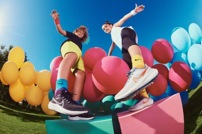 Two kids strike playful poses beside balloons and athletic shoes. Nike shoes are featured on a platform against a blue sky.