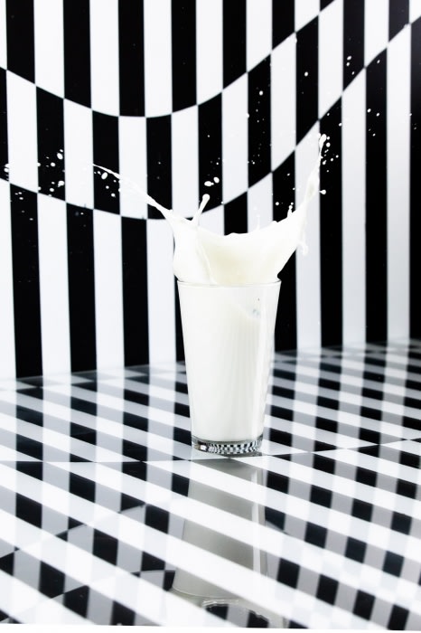 A glass of milk creates a splash against a black and white geometric background, emphasizing texture, color contrast, and fluid motion.