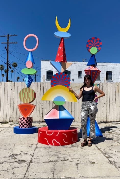 Young woman stands in front of two colorful abstract art sculptures against a white wooden fence and blue sky. Sunny day.