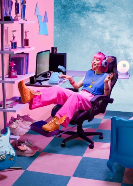 Woman with pink hair and headset at a computer desk in a colorful interior room with a computer setup, chair, and decor.