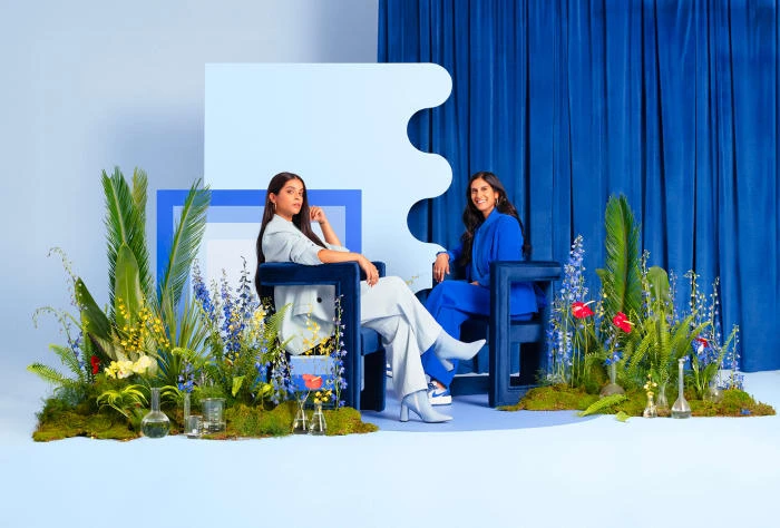 Two women in blue and white suits sit in blocky blue chairs surrounded by lush floral arrangements against a blue and white backdrop.