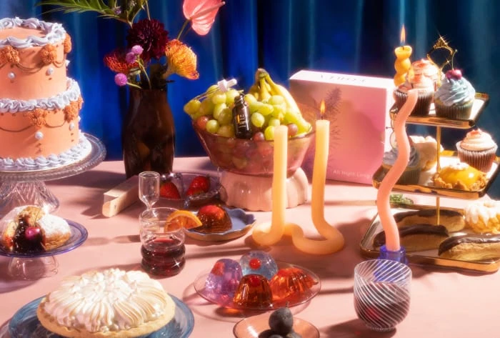 Table arrangement featuring a two-tiered cake, assortment of desserts, fruit bowl, pink box, lighted candles against blue curtains.