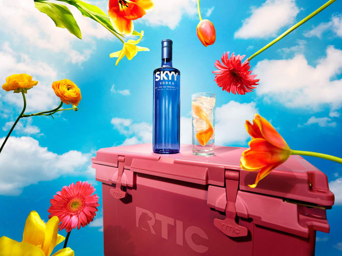 Skyy Vodka bottle and cocktail glass on pink cooler against a cloudy blue sky with colorful flowers in a commercial still life.