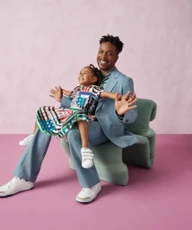 Black man in a light blue suit holding a smiling young girl in a patterned colorful dress seated on a mint green chair