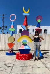Set designer Gabriela Cobar posing beside colourful geometric sculptures outdoors on an asphalt lot