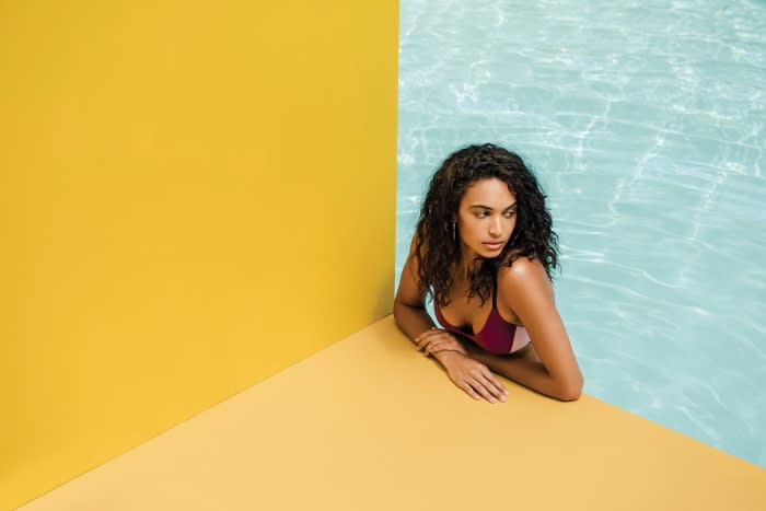 Woman in swimwear poses near the corner of a swimming pool against a yellow wall, showcasing a blend of fashion and leisure.