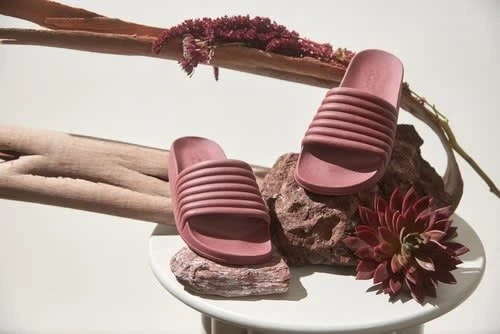 Two slide sandals in a mauve color are displayed on rocks and a small white table with decorative botanicals against a white background.