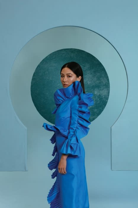 Elegant woman in a blue ruffled gown posing against a light blue backdrop with a geometric circular pattern in a fashion photography studio.