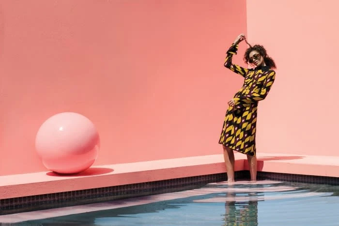Woman in patterned dress stands in pool, pink wall and round ball background. Fashion shot with vibrant colors and unique setting.