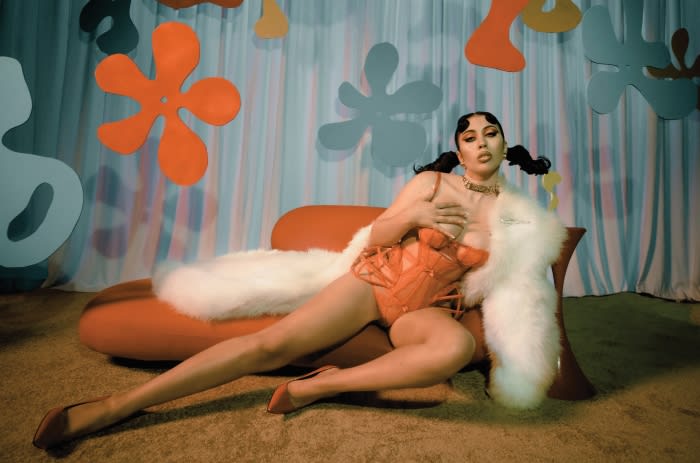 Glamorous adult female reclining on an orange sofa in a coral bodysuit and white boa in front of a retro-styled backdrop.