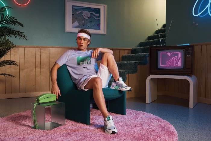 Man in lilac Puma shirt and white shorts lounges in green chair, with MTV on TV, retro interior, lit by neon lights.