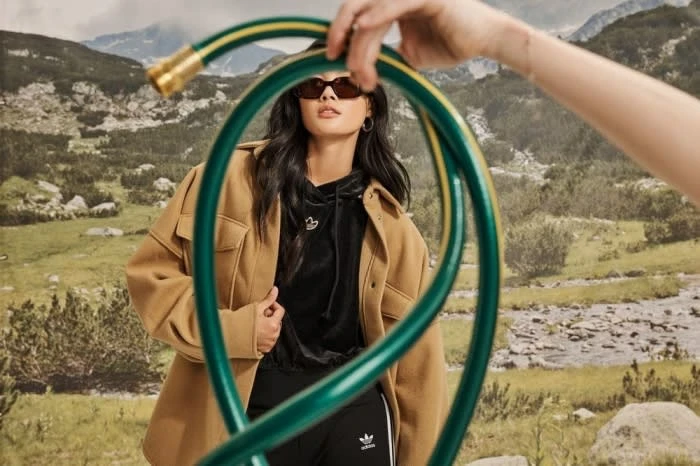 Woman wearing sunglasses and jacket stands near a garden hose outdoors with mountains, plants, and sky visible in the background.