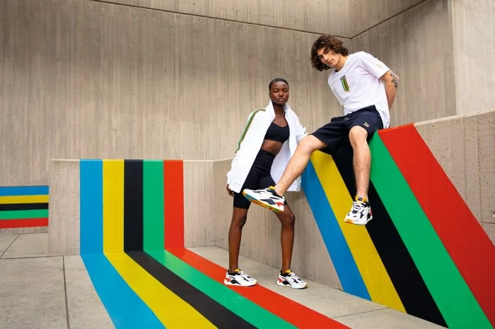 Two fashion models pose in PUMA athletic wear, including sneakers, against a colorful striped wall and concrete architectural background.