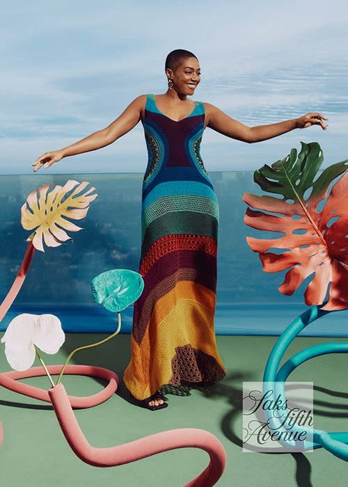 Woman in a colorful crochet dress stands in a studio with tropical-themed decor elements, showcasing a blend of fashion and art.