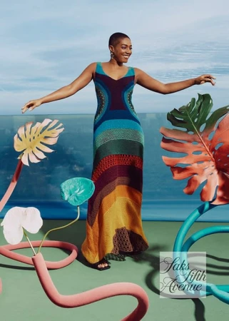 Woman in a colorful crochet dress stands in a studio with tropical-themed decor elements, showcasing a blend of fashion and art.