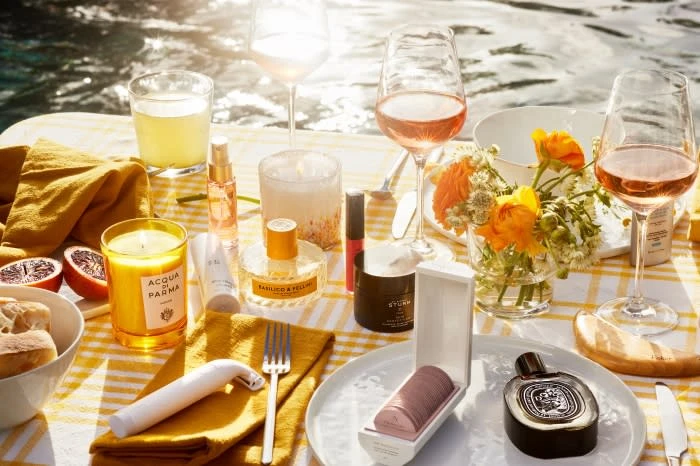 Still life of perfume bottles, candles, beverages and flowers arranged on a yellow gingham tablecloth near water, bright sunlight.