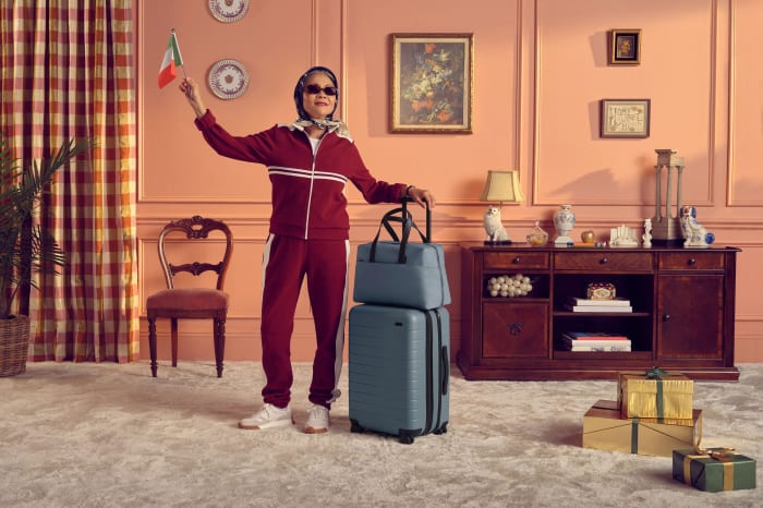 Woman in tracksuit waves Italian flag indoors, standing beside luggage and gift boxes with peach-colored walls and decorative details.