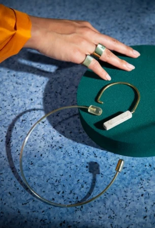 Hand with gold rings resting near modern metal jewelry pieces on a green platform and speckled blue surface.