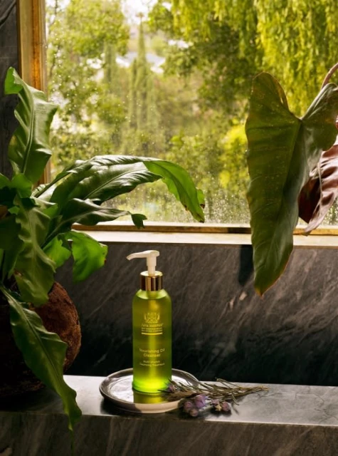 An eye-level shot of a bottle of cleansing oil on a marble surface, adorned with plants and flowers, with a bright outdoor background.