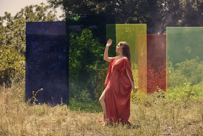 Woman in red dress poses in golden field against backdrop of multicolored transparent screens, creating vibrant play of color and light.