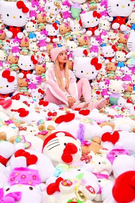Young woman in a pink outfit sits amongst many Hello Kitty plush toys; bright, fun, vibrant and colorful scene with pink color dominance.