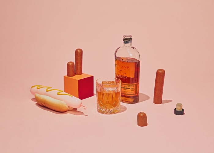 Still life on pink backdrop featuring hot dog with mustard, glass of whiskey, bottle, sausage pieces, and orange and red podium.