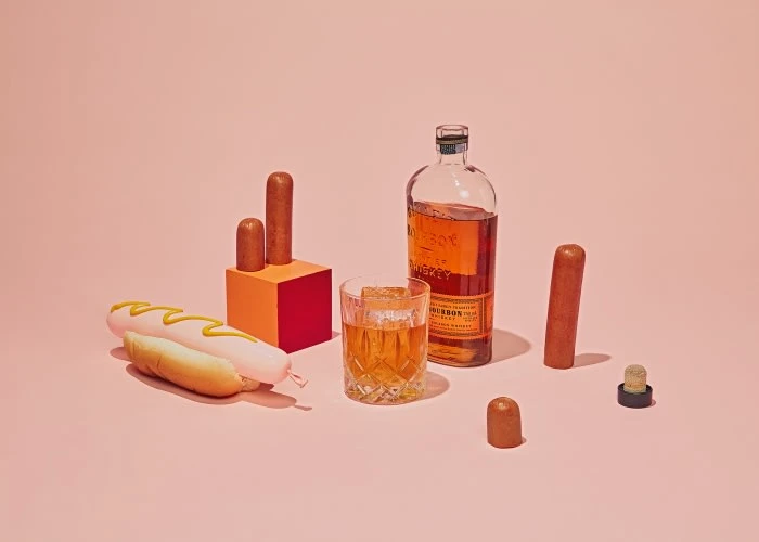 Still life on pink backdrop featuring hot dog with mustard, glass of whiskey, bottle, sausage pieces, and orange and red podium.