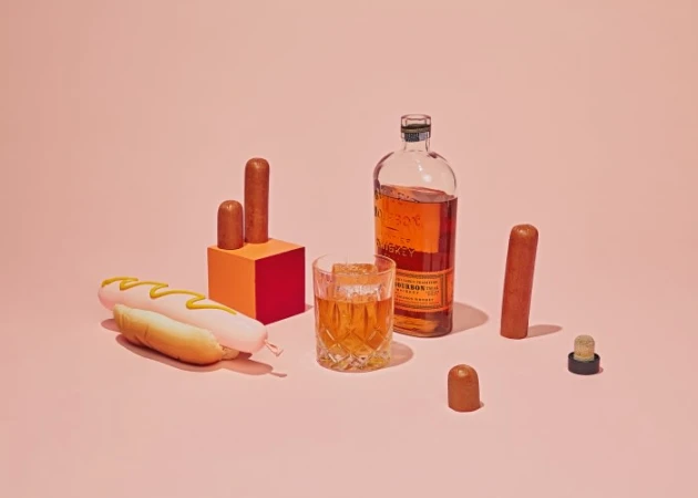 Still life on pink backdrop featuring hot dog with mustard, glass of whiskey, bottle, sausage pieces, and orange and red podium.