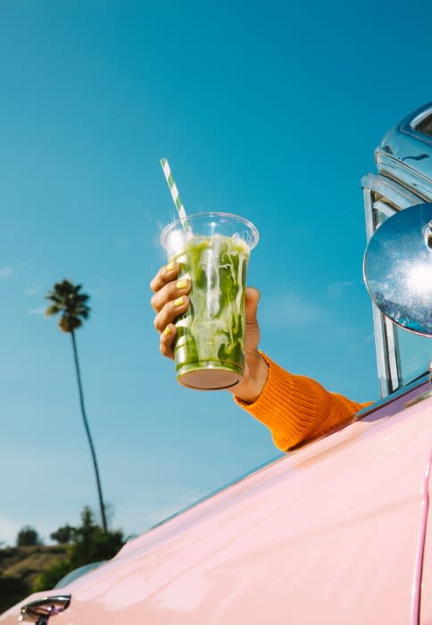 A refreshing green smoothie with a striped straw is held out of a vintage pink car under a clear blue sky with a palm tree.