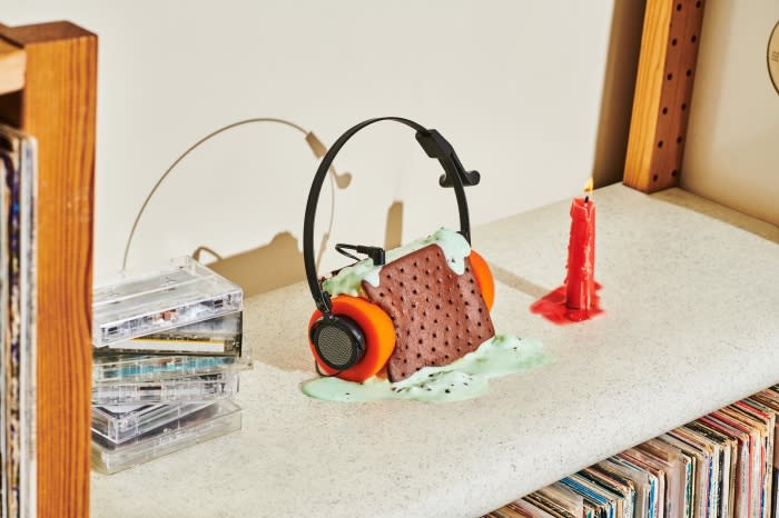 Headphones with ice cream sandwich on shelf with records, audio cassettes, and a lit candle create an unusual visual.