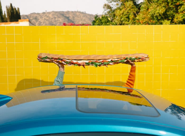 Extra-long sandwich held above a blue car, contrasting with a vibrant yellow tiled wall and a backdrop of lush trees and hills.