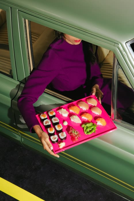 Woman in a green vintage car holds a tray of sushi. The scene features an indoor shot with a yellow stripe marking on the asphalt.