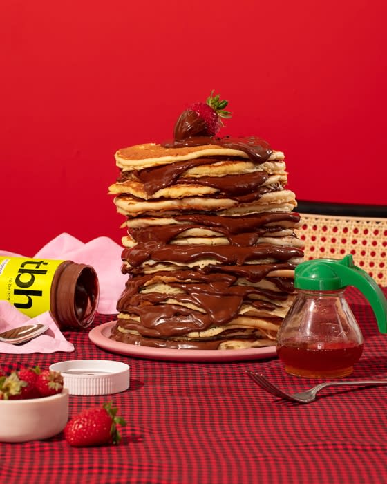 Stack of pancakes with chocolate sauce and a strawberry on top, accompanied by a jar of honey, atop a red checkered tablecloth.