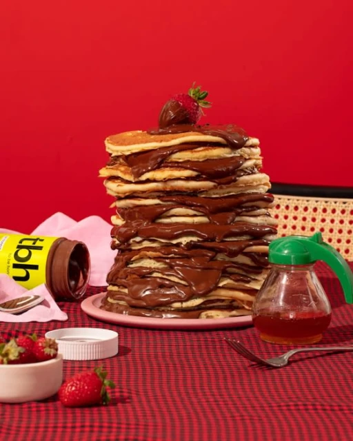 Stack of pancakes with chocolate sauce and a strawberry on top, accompanied by a jar of honey, atop a red checkered tablecloth.