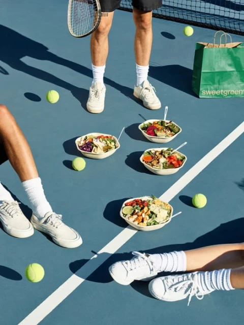 Overhead shot of salads and tennis balls on a tennis court, with three partial people visible and Sweetgreen bag in background.