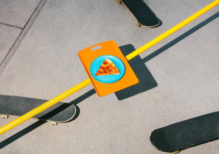 Eye-level shot featuring a slice of pizza on a plate, balanced on a cutting board placed across a yellow metal bar over a concrete floor with skateboards.