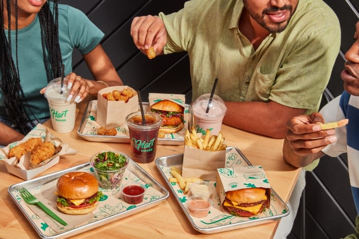 A group enjoys plant-based fast food at Hart House, featuring burgers, nuggets, fries, and beverages on metal trays on a wooden table.