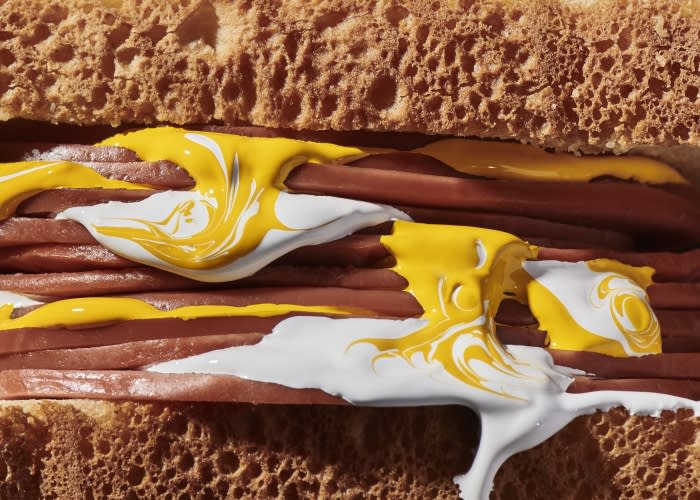 Overhead close-up of a gourmet hot dog on a toasted bun, smothered in creamy white and yellow sauces, highlighting textures.