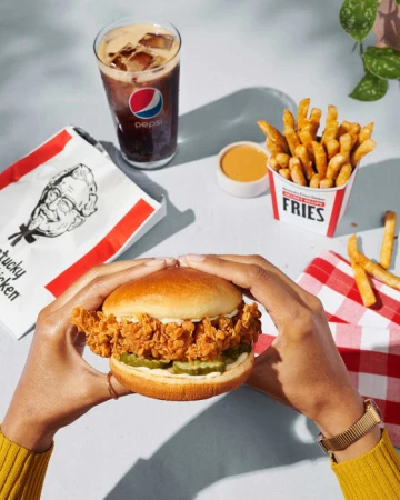 Crispy chicken sandwich held by hands, with a side of french fries, dipping sauce, and a Pepsi drink. Food photography.