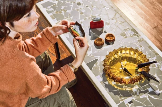 Woman prepares roll-up with lighter, grinder, and amber ashtray on mosaic table indoors under natural lighting; overhead, angled shot.