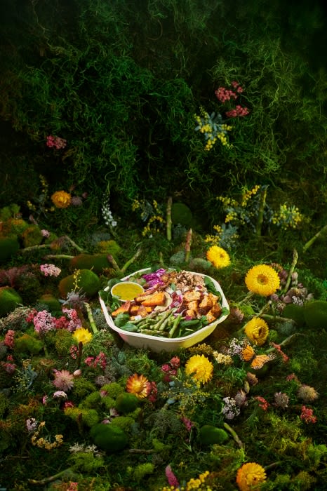 Bowl of vibrant salad with grilled chicken, asparagus, and mixed greens on a bed of moss, surrounded by flowers and foliage.