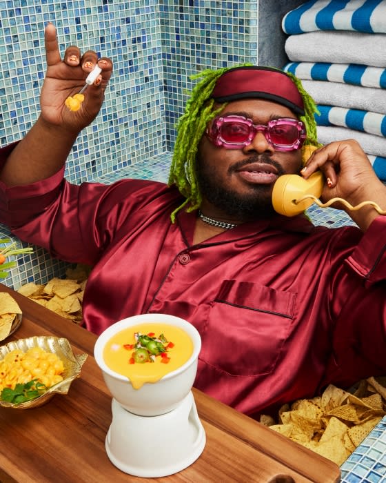 Person in burgundy pajamas reclines in a bathtub of tortilla chips, talking on a yellow phone, with cheese dip and mac and cheese.