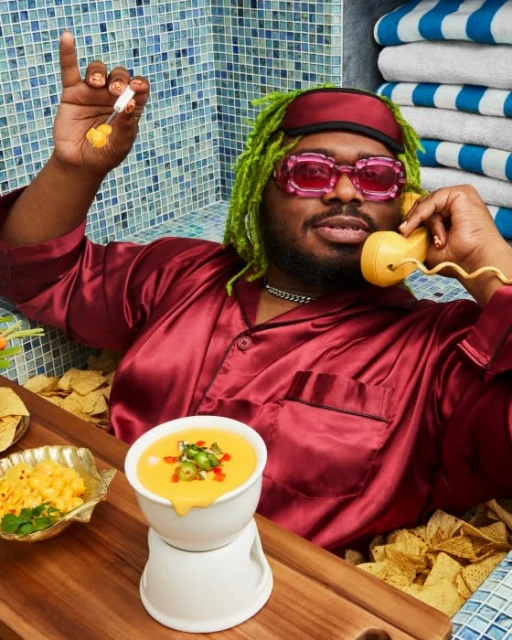 Person in burgundy pajamas reclines in a bathtub of tortilla chips, talking on a yellow phone, with cheese dip and mac and cheese.