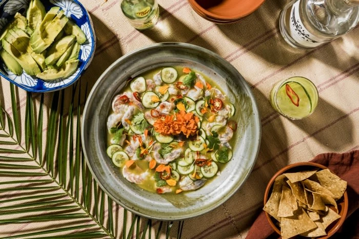 Overhead shot of ceviche in a bowl surrounded by avocado slices, tortilla chips, and a cocktail on a striped tablecloth.
