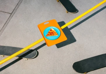 A slice of pepperoni pizza on a blue plate atop an orange tray, bisected by a long yellow rail on a grey concrete surface with skateboards.